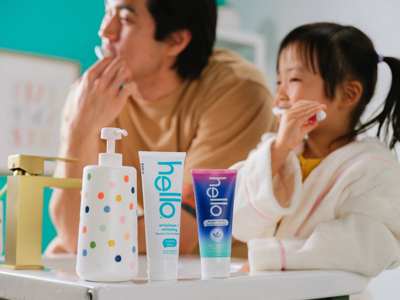 Father and daughter brushing teeth with 'hello' toothpaste and products on a bathroom counter.